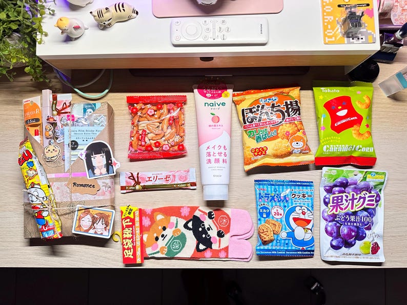 Puede incluir: Una colecci&oacute;n de snacks, productos para el cuidado de la piel y art&iacute;culos de papeler&iacute;a japoneses dispuestos sobre una superficie de madera. Incluye bolsas de patatas fritas, un tubo de limpiador facial y un paquete de galletas. Tambi&eacute;n se ven pegatinas decorativas y un par de calcetines Shiba Inu.