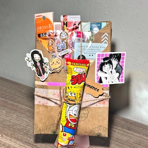 Puede incluir: Una caja de regalo de papel marr&oacute;n decorada con pegatinas, cinta washi y una barra de caramelo amarilla con un personaje de dibujos animados. La caja est&aacute; atada con hilo y lleva una etiqueta peque&ntilde;a que dice "romance".