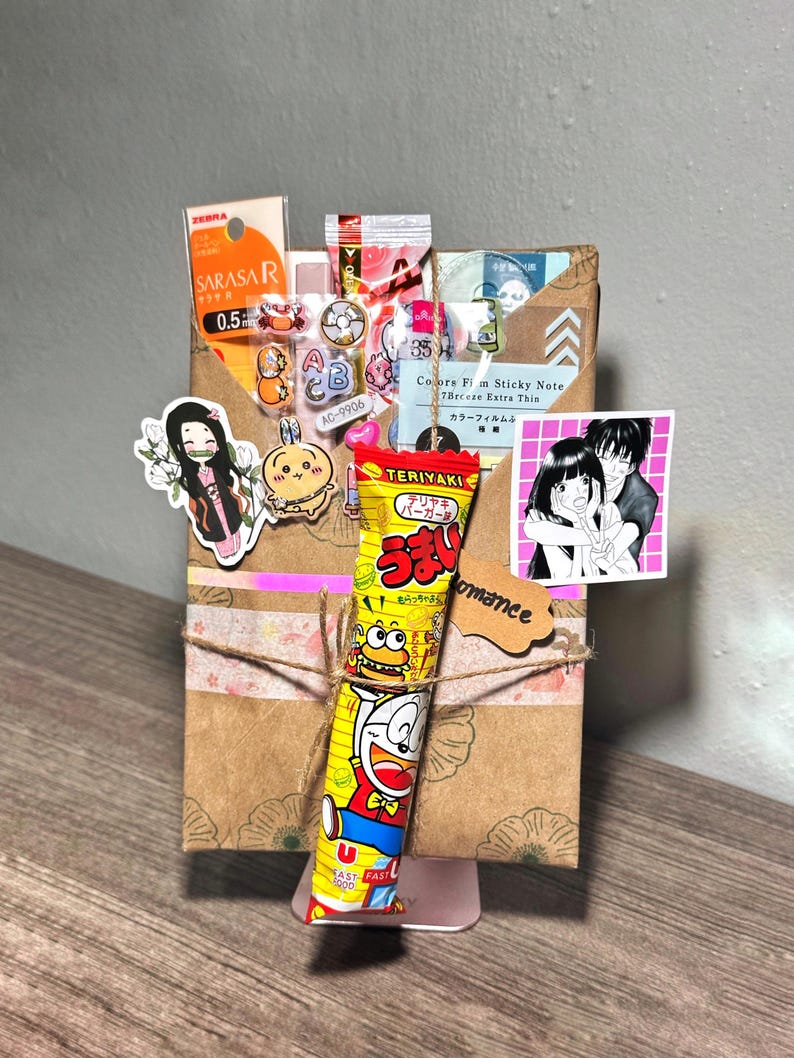 Puede incluir: Una caja de regalo de papel marr&oacute;n decorada con pegatinas, cinta washi y una barra de caramelo amarilla con un personaje de dibujos animados. La caja est&aacute; atada con hilo y lleva una etiqueta peque&ntilde;a que dice "romance".