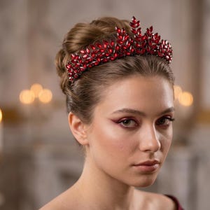 Op de afbeelding: Een vrouw draagt een rode, met juwelen bezette tiara. De tiara is gemaakt van rode edelstenen en is gezet op een zilverkleurige metalen basis. De vrouw heeft haar haar in een knot. De achtergrond is wazig, met kaarsen en een muur zichtbaar.