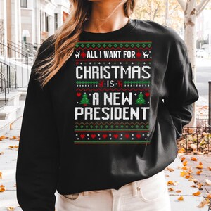 May include: Black sweatshirt with a festive Christmas design. The text reads "ALL I WANT FOR CHRISTMAS IS A NEW PRESIDENT" in white, green, and red, with reindeer, Christmas trees, and heart patterns.