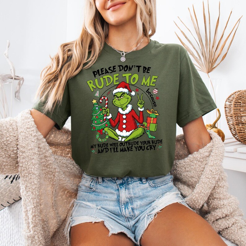 May include: Olive green t-shirt featuring a Grinch character in a Santa hat and red suit, holding a candy cane and Christmas tree. Text reads "PLEASE DON'T BE RUDE TO ME" and "MY RUDE WILL OUTRUDE YOUR RUDE AND I'LL MAKE YOU CRY".