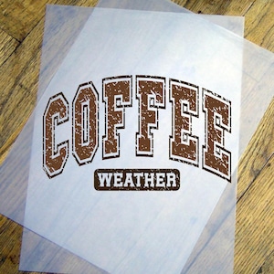 May include: A sheet of translucent material with the words "COFFEE WEATHER" printed in brown, collegiate-style lettering. The text is centered on the sheet, which is placed on a wooden surface.
