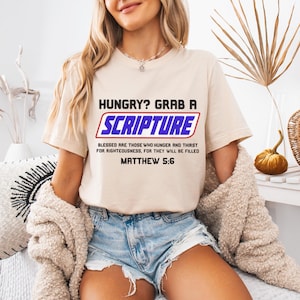 May include: Beige t-shirt with the text "HUNGRY? GRAB A SCRIPTURE" in black and blue. Below the text is the quote "BLESSED ARE THOSE WHO HUNGER AND THIRST FOR RIGHTEOUSNESS, FOR THEY WILL BE FILLED" and "MATTHEW 5:6".