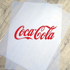 May include: Two sheets of translucent paper with the red Coca-Cola logo printed on the top sheet. The logo is in the classic script font. The paper is on a light brown wooden surface.