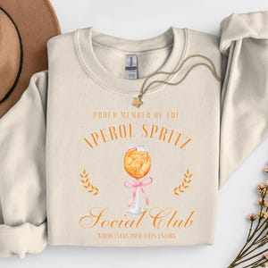 May include: Cream-colored sweatshirt with the text "PROUD MEMBER OF THE APEROL SPRITZ SOCIAL CLUB" and a watercolor illustration of an Aperol Spritz cocktail. A gold necklace with a star pendant is draped on the sweatshirt.