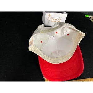 Op de afbeelding: Een witte baseballpet met een rode klep, van binnenuit bekeken. De pet heeft een witte verstelbare band en rode stiksels. De klep is felrood. De pet is waarschijnlijk gemaakt van een katoen- of polyestermix.