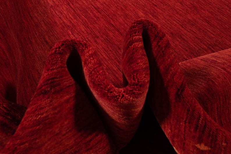 Pu&ograve; includere: Primo piano di un tappeto rosso piegato con una superficie strutturata. Le fibre del tappeto creano un motivo di linee e ombre, con una tonalit&agrave; rosso intenso. Le pieghe aggiungono profondit&agrave; e dimensione al tessuto.