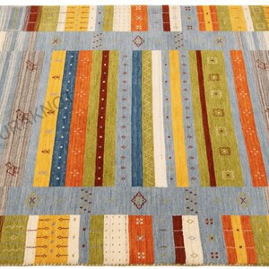 May include: A rectangular area rug with a light blue central field and colorful vertical stripes in orange, yellow, green, and red. The rug has a cream-colored border with geometric patterns and a diamond design.