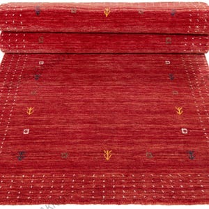 Op de afbeelding: Een rode vloerkleed met een geometrisch patroon. Het kleed is aan de bovenkant opgerold, waardoor de onderkant zichtbaar is. Het kleed heeft een rand van kleine witte stippen en is versierd met kleine, kleurrijke geometrische vormen.