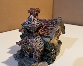 May include: A miniature decorative house with a stone facade, a red-tiled roof, and a chimney. The house has a small door and windows, with green shrubbery around the base. The word "BOYDS" is above the door.