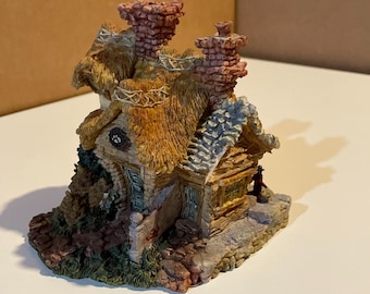 May include: A miniature cottage figurine with a thatched roof, brick chimneys, and stone walls. The cottage has a small door and windows, surrounded by greenery. The color palette includes shades of brown, beige, and green, creating a rustic aesthetic.