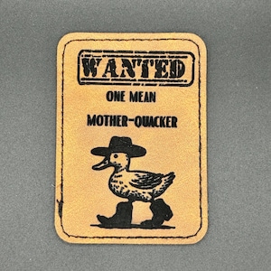 Peut inclure: Un écusson rectangulaire de couleur beige avec une bordure noire. L'écusson présente le mot "WANTED" dans un cadre rectangulaire en haut, avec le texte "ONE MEAN MOTHER-QUACKER" en dessous. Un canard de dessin animé portant un chapeau et des bottes est au centre.