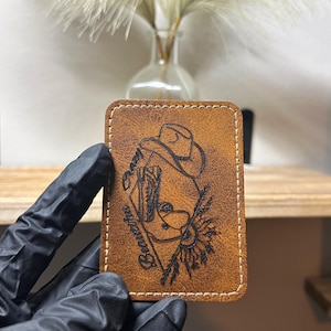 May include: A rectangular, brown leather patch with a stitched border. The patch features a detailed, etched design of a cowboy wearing a hat, with the word "Beautiful" and a sunflower. The patch is held by a gloved hand.