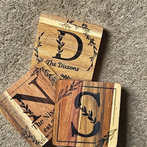 Może przedstawiać: Trzy drewniane kwadratowe podstawki z personalizowanymi wzorami liter. Każda podstawka ma inną literę, z wygrawerowanymi pod spodem imionami "The Dixsons" i "The Nixs". Litery otoczone są ozdobnym wzorem liści.