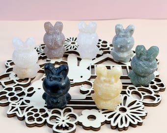 May include: A collection of seven carved stone bunny figurines in various colors, including white, gray, and yellow. The bunnies are arranged on a decorative wooden base with a floral and bunny design. The background is a soft pink.