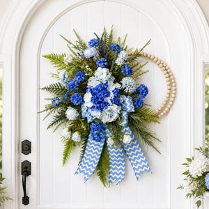 May include: A teardrop-shaped floral wreath featuring blue and white hydrangeas, green fern fronds, and a natural wood bead accent. A blue and white chevron patterned ribbon hangs from the center, adding a geometric element to the arrangement.