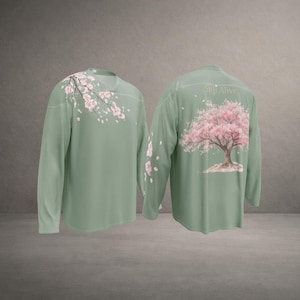 May include: Sage green long-sleeve shirt featuring a cherry blossom design. The front of the shirt has a branch of pink flowers, while the back displays a full cherry blossom tree. The text "Still Alive" is above the tree.
