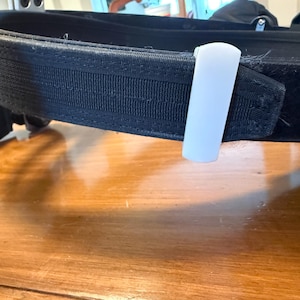 May include: A black tactical utility belt featuring a thick, durable nylon strap with reinforced stitching and a smooth, white plastic belt keeper loop attached to the exterior. The belt has a textured surface and includes black hardware components.
