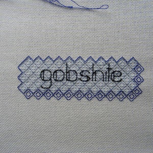 May include: A close-up of a white fabric with blue stitching. The word "gobsh*te" is stitched in black thread.