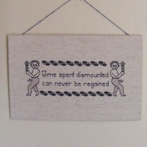 May include: A beige canvas wall hanging with black embroidery. The embroidery depicts two figures holding swords and the text "Time spent dismounted can never be regained."