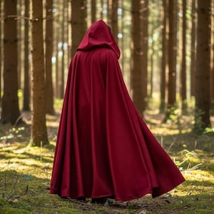 May include: A long, flowing, burgundy hooded cloak. The cloak is made of a soft fabric and is worn in a forest setting. The hood is up, and the cloak drapes down to the ground. The background is a forest with tall trees and sunlight.