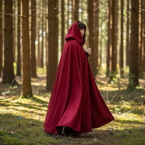 May include: A long, flowing, crimson hooded cloak. The cloak is worn in a forest setting with tall trees and dappled sunlight. The garment is made of a heavy fabric and drapes to the ground.
