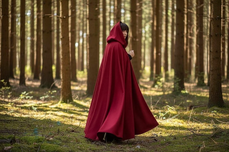 May include: A long, flowing, crimson hooded cloak. The cloak is worn in a forest setting with tall trees and dappled sunlight. The garment is made of a heavy fabric and drapes to the ground.