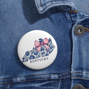 Puede incluir: Un botón blanco con el contorno de Kentucky, relleno con un estampado floral azul. Un lazo rosa está centrado sobre el contorno del estado. La palabra "KENTUCKY" está impresa en azul marino debajo.