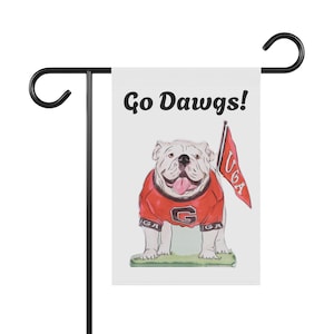 May include: A white garden flag featuring a cartoon bulldog wearing a red jersey with a black G logo and the text Go Dawgs! at the top. The bulldog holds a red pennant flag with the letters UGA in white, standing on a small patch of green grass.