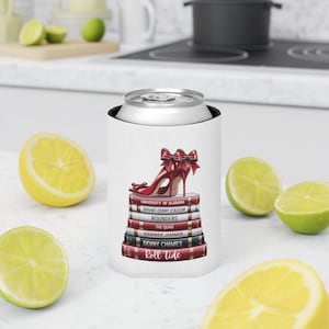 May include: A white can cooler with a red high-heeled shoe and bow design, placed on a stack of books. The books are labelled with titles such as "Roll Tide" and "University of Alabama." The design incorporates red, white, and black. Lemons and limes are in the background.
