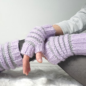 Puede incluir: Un par de guantes sin dedos de color lavanda con rayas grises. Los guantes de punto están hechos de un material suave y están diseñados para mantener las manos calientes mientras dejan los dedos libres. Los guantes se muestran sobre un fondo blanco.