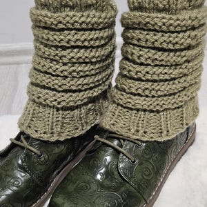 Puede incluir: Polainas de punto verde oliva sobre zapatos estampados verde oscuro. Las polainas tienen un puño acanalado y un diseño apilado. Los zapatos tienen un acabado brillante y un patrón de remolino decorativo. Las polainas miden unos 20 cm de largo.