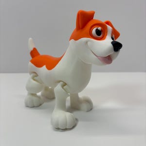 May include: A cartoon-style dog figurine with a white body and orange markings on its head and back. The dog has a black nose, big eyes, and a smiling expression. The figurine is standing on all fours.