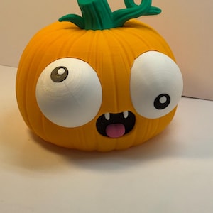 May include: A bright orange pumpkin figurine with a green stem, featuring two large white eyes with black pupils, a black mouth with two white fangs, and a small pink tongue sticking out. The pumpkin has a ribbed texture and a rounded shape.