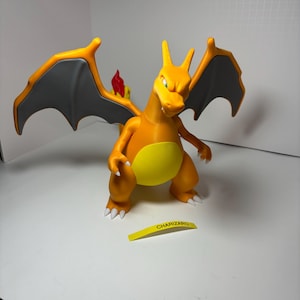 May include: A 3D-printed orange Charizard figure with gray wings and a yellow belly. The figure has a flame on its tail and a yellow label that reads "CHARIZARD". The figure is standing on a white surface.