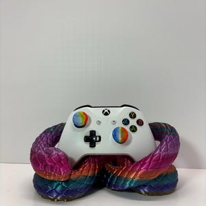 May include: White Xbox controller with rainbow-colored buttons and a decorative, multi-colored snake base. The snake base features shades of purple, pink, blue, and gold, creating a vibrant contrast with the controller.