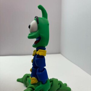 Puede incluir: Una figura de acci&oacute;n verde articulada con un ojo blanco grande, detalles azules y amarillos y una boca sonriente. La figura tiene un cuerpo de gusano y est&aacute; posada sobre una superficie blanca. La figura es un personaje de dibujos animados.