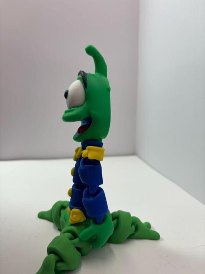 Puede incluir: Una figura de acci&oacute;n verde articulada con un ojo blanco grande, detalles azules y amarillos y una boca sonriente. La figura tiene un cuerpo de gusano y est&aacute; posada sobre una superficie blanca. La figura es un personaje de dibujos animados.