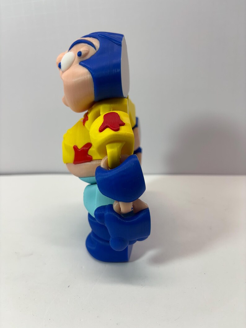 K&ouml;nnte beinhalten: 3D-gedruckte Actionfigur einer Zeichentrickfigur. Die Figur hat einen blauen Helm, einen gelben Torso und blaue Stiefel. Rote Akzente befinden sich auf den Schultern und der Brust. Die Figur steht auf einer wei&szlig;en Oberfl&auml;che.