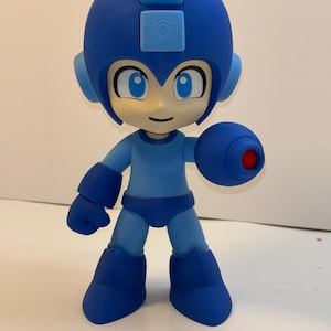 May include: A blue action figure of a character with a helmet, large boots, and a cannon arm. The figure has a light blue face with blue eyes and a small smile. The figure is standing on a white surface.