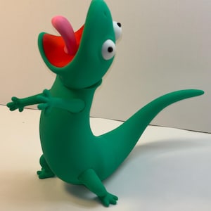 May include: A green cartoon chameleon figurine with a pink tongue extended. The chameleon has large, white-and-black eyes and is posed with its arms outstretched. The inside of its mouth is red.