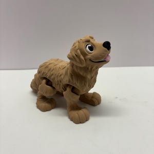 May include: A light brown, articulated toy dog with a friendly expression. The dog has a black nose, white eyes, and a pink tongue sticking out. The toy is designed to be poseable, with movable legs and a textured fur appearance.