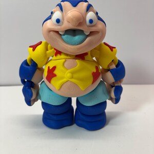 K&ouml;nnte beinhalten: Eine farbenfrohe Figur einer Zeichentrickfigur mit gro&szlig;em Kopf, blauen Haaren und einer herausragenden Zunge. Die Figur tr&auml;gt ein gelbes Hemd mit roten Akzenten, blaue Arm- und Beinbedeckungen und hellblaue Shorts. Die Figur ist etwa 13 cm gro&szlig;.