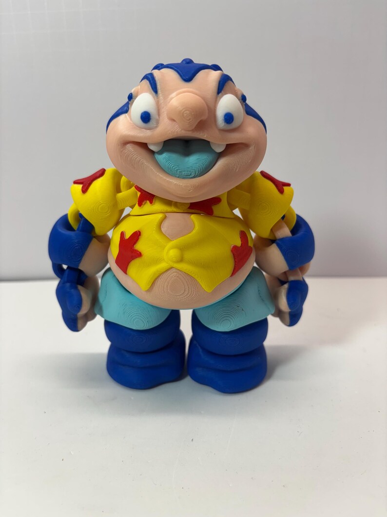 K&ouml;nnte beinhalten: Eine farbenfrohe Figur einer Zeichentrickfigur mit gro&szlig;em Kopf, blauen Haaren und herausgestreckter Zunge. Die Figur tr&auml;gt ein gelbes Hemd mit roten Akzenten, blaue Arm- und Beinbedeckungen und hellblaue Shorts. Die Figur steht aufrecht.