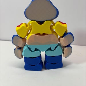 K&ouml;nnte beinhalten: Ein farbenfroher, beweglicher Spielzeugroboter mit blauem Kopf, gelbem Torso und blauen Beinen. Die Arme des Roboters sind gelb mit roten Akzenten. Der Mittelteil ist pfirsichfarben und hellblau. Das Spielzeug ist mit mehreren Gelenken versehen.