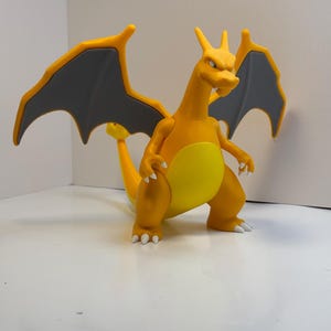 May include: A plastic Charizard figure, featuring a yellow belly, orange body, and gray wings. The dragon figure has white claws and a long tail. The wings are spread, and the figure is posed in a dynamic stance.
