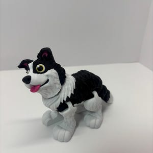 May include: A black and white Border Collie figurine with a pink tongue and yellow eyes. The dog is in a playful pose, with its mouth open and tongue sticking out. The figurine has a fluffy white chest and legs.