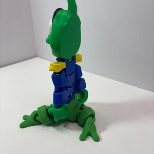 Puede incluir: Una figura articulada verde y azul con una cabeza grande y una antena curva. La figura tiene detalles amarillos en los hombros y est&aacute; en posici&oacute;n sentada. Las extremidades de la figura est&aacute;n hechas de segmentos conectados.