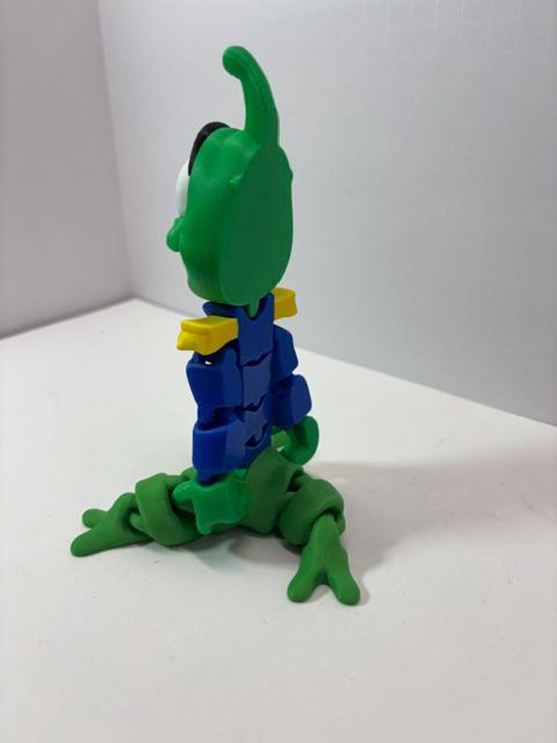 Puede incluir: Una figura articulada verde y azul con una cabeza grande y una antena curva. La figura tiene detalles amarillos en los hombros y est&aacute; en posici&oacute;n sentada. Las extremidades de la figura est&aacute;n hechas de segmentos conectados.